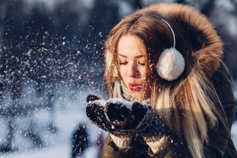 Eine Person mit langen Haaren, die einen Wintermantel mit pelzgefütterter Kapuze und Ohrenschützern trägt, bläst in einer verschneiten Außenumgebung Schnee aus ihren Händen. Der Hintergrund ist verschwommen und zeigt Silhouetten von Menschen und Bäumen.