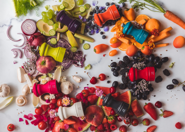 Eine Draufsicht auf verschiedene Obst- und Gemüsesorten, die nach Farben auf einer Marmoroberfläche angeordnet sind. Sechs bunte Mühlen sind zwischen den Produkten platziert, jede davon passend zu den Lebensmittelfarben um sie herum, wodurch eine lebendige und künstlerische Präsentation entsteht.