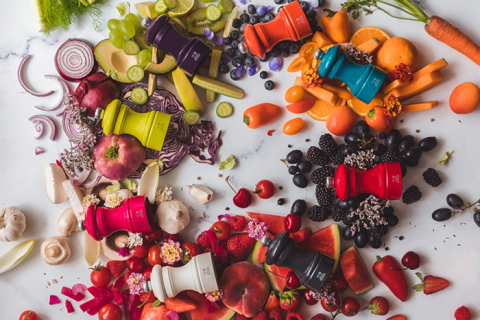 Eine Draufsicht auf verschiedene Obst- und Gemüsesorten, die nach Farben auf einer Marmoroberfläche angeordnet sind. Sechs bunte Mühlen sind zwischen den Produkten platziert, jede davon passend zu den Lebensmittelfarben um sie herum, wodurch eine lebendige und künstlerische Präsentation entsteht.