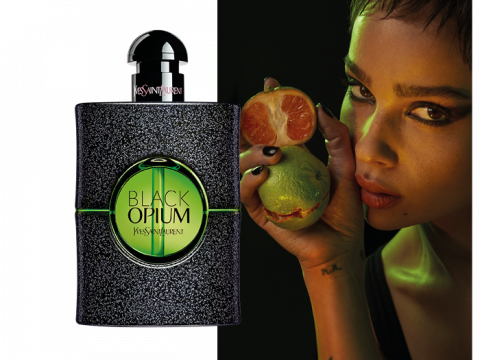 Eine Frau mit kurzen Haaren hält eine geschälte Zitrusfrucht vor ihr Gesicht. Neben ihr steht eine Flasche Yves Saint Laurent Black Opium Eau de Parfum mit einem dunklen Glitzerdesign und leuchtend grünen Akzenten. Der Hintergrund ist schwach beleuchtet.