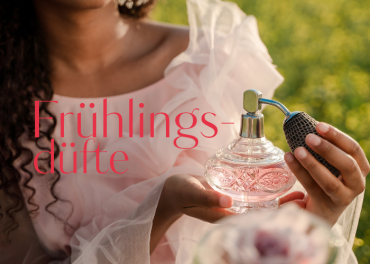 Eine Person in einem zartrosa Rüschenkleid hält eine verzierte rosa Parfümflasche mit einem Zerstäuberkolben. Der Text „Frühlingsdüfte“ ist in Rot zu sehen. Der Hintergrund ist mit üppigem Grün gestaltet.