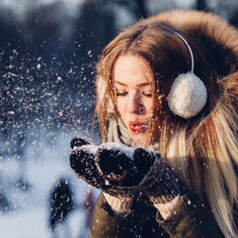 Eine Person in Wintermantel und Ohrenschützern bläst Schnee aus ihren behandschuhten Händen und erzeugt eine Wolke aus Schneeflocken. Der Hintergrund zeigt eine verschneite Landschaft und verschwommene Menschen, was eine kühle, fröhliche Winterszene vermittelt.