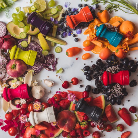 Bunte Pfeffermühlen sind auf einer Marmoroberfläche angeordnet, jede von einer Vielfalt passender Früchte und Gemüsesorten wie Zitrusfrüchten, Beeren und Paprika umgeben, wodurch eine lebendige und frische Präsentation entsteht.