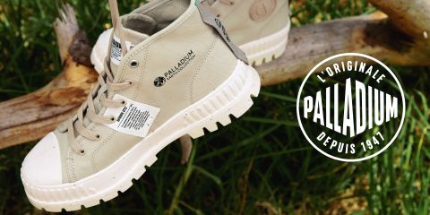 Ein Paar beige Palladium High-Top-Sneaker hängt draußen an einem Ast. Der Schuh hat eine weiße Sohle und ein Etikett mit Text. Auf der rechten Seite steht auf einem runden Logo „L'Originale Palladium Depuis 1947“. Im Hintergrund ist Gras zu sehen.