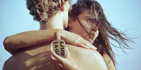 Eine Frau mit nassen Haaren umarmt einen Mann am Strand von hinten. Sie hält ihm eine Glasflasche mit Korken mit der Aufschrift „O SUS“ auf den Rücken. Im Hintergrund ist der Himmel klar.
