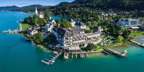 Luftaufnahme eines Resorts am Seeufer mit mehreren Gebäuden, umgeben von üppigem Grün. Das Resort liegt auf einer kleinen Halbinsel mit Anlegestellen und Booten, neben türkisfarbenem Wasser und sanften Hügeln unter einem klaren Himmel.