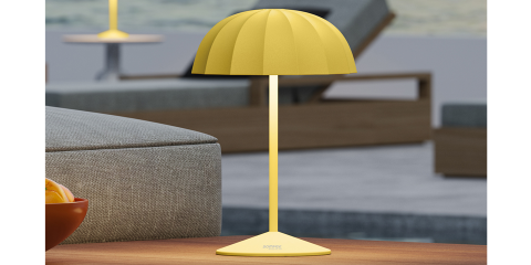Eine elegante gelbe Tischlampe mit kuppelförmigem Schirm steht auf einem Holztisch. Das minimalistische Design der Lampe ergänzt die modernen Gartenmöbel im unscharfen Hintergrund.