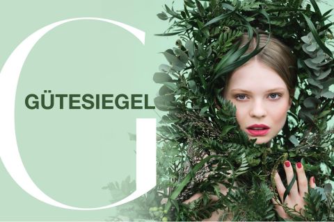 Eine Frau mit leuchtend rotem Lippenstift ist von üppigem grünem Laub vor einem hellgrünen Hintergrund umgeben. Das Wort „GÜTESIEGEL“ steht neben einem großen Buchstaben „G“ auf der linken Seite des Bildes.