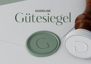 Ein Wachssiegel mit dem Buchstaben „G“ und dem Wort „Gütesiegel“ darüber auf weißem Untergrund, neben einem Stempel. Der Hintergrund ist hell und der Stempelgriff hat eine Holzstruktur.