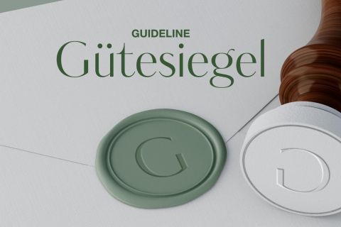 Ein Wachssiegel mit dem Buchstaben „G“ und dem Wort „Gütesiegel“ darüber auf weißem Untergrund, neben einem Stempel. Der Hintergrund ist hell und der Stempelgriff hat eine Holzstruktur.