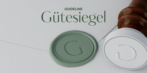 Abgebildet ist ein Holzstempel mit einem grünen Wachssiegelabdruck mit dem Buchstaben „G“. Darüber steht in grüner Schrift das Wort „Gütesiegel“. Die Szene suggeriert ein Thema der Qualitätssicherung oder Zertifizierung.