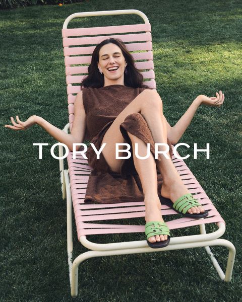 Eine lächelnde Frau in einem braunen Kleid entspannt sich mit ausgebreiteten Armen und grünen Sandalen im Freien auf einem rosa Liegestuhl. Der Text „TORY BURCH“ ist in der Bildmitte eingeblendet.