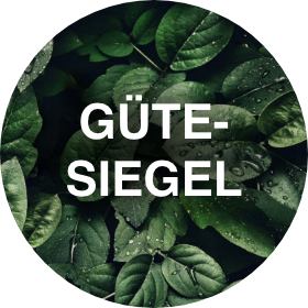Ein kreisförmiges Bild mit üppigen grünen Blättern, die mit Wassertropfen bedeckt sind, als Hintergrund. Der Text „GÜTESIEGEL“ ist in der Mitte deutlich in weißen Großbuchstaben zu sehen.