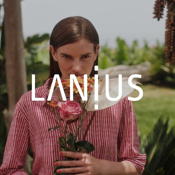 Eine Person, die Blumen vor ihr Gesicht hält, steht im Freien, umgeben von Grün. Sie trägt ein rot-weiß gestreiftes Hemd. Das Wort „LANIUS“ ist über das Bild gelegt.
