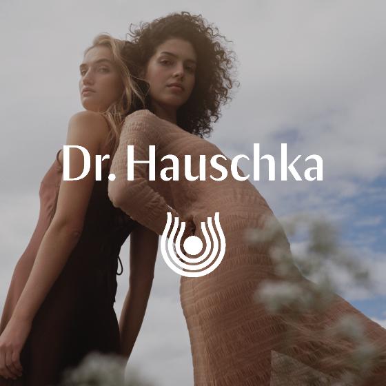 Zwei Frauen in eleganten Kleidern posieren vor einem wolkigen Himmel. Das Dr. Hauschka Logo und der Text sind über das Bild gelegt.