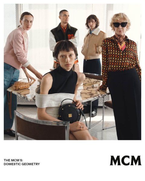 Fünf stilvolle Personen posieren in einem Innenraum um einen Marmortisch herum und präsentieren MCM-Taschen und Mode in einem modernen, eleganten Ambiente.