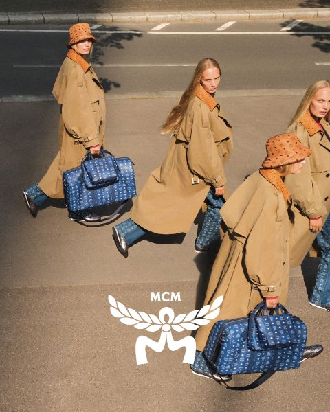 Drei Frauen in hellbraunen Mänteln und orangefarbenen Hüten gehen über eine Straße und tragen blaue MCM-Taschen mit einem weißen MCM-Logo im Vordergrund. Das Bild hat einen Spiegeleffekt, der die Szene dupliziert erscheinen lässt.