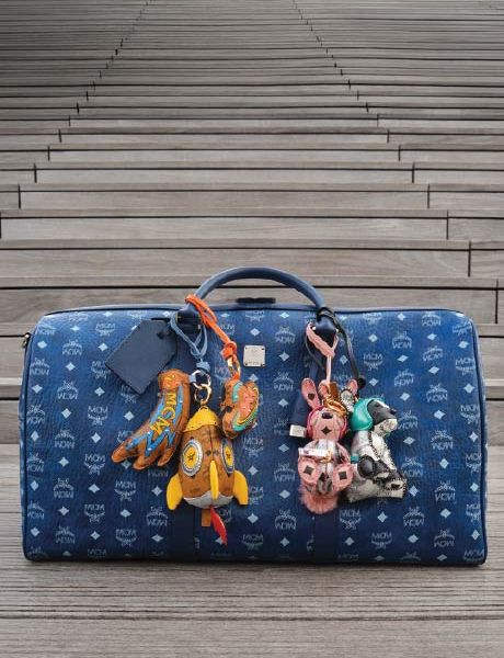 Eine blaue MCM-Designertasche mit weißem Logomuster steht auf hölzernen Stufen, die mit bunten Tier- und Objektschlüsselbändern geschmückt sind, darunter eine Rakete, ein Hase und ein Bär.