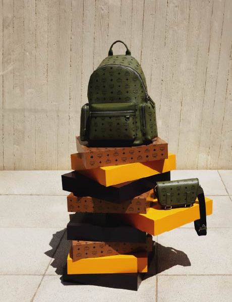 Ein grüner Designerrucksack und eine dazu passende Umhängetasche stehen auf einem Stapel orangefarbener und brauner Kisten vor einer beigefarbenen Strukturwand und einem gefliesten Boden.