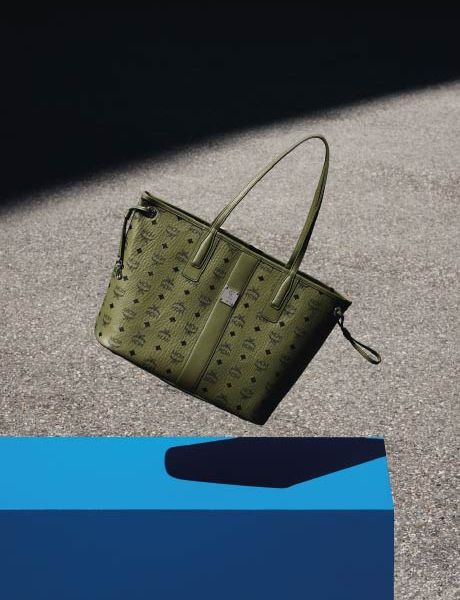 Eine grüne Designer-Handtasche mit dezentem Monogrammmuster ruht auf einer grauen Fläche, mit einem blauen geometrischen Objekt im unteren Vordergrund und einem Schattenwurf im Hintergrund.