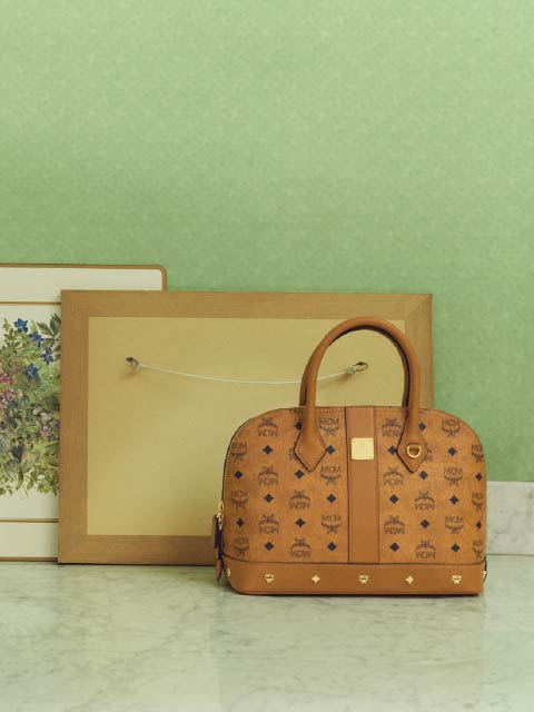 Eine braune Designerhandtasche liegt auf einer Marmorfläche vor zwei gerahmten Bildern vor einer hellgrünen Wand.