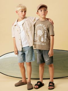 Zwei junge Jungen stehen nebeneinander vor einem beigen Hintergrund. Einer trägt ein hellbraunes Hemd, ein weißes T-Shirt und Jeansshorts, während der andere eine braune Mütze, ein bedrucktes T-Shirt und dunkle Shorts trägt. Hinter ihnen ist ein Surfbrett zu sehen.