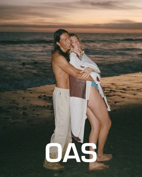 Ein Paar umarmt sich bei Sonnenuntergang an einem Strand. Der Mann ist oben ohne und trägt leichte Hosen, während die Frau in einen weiß-braunen Schal gehüllt ist. Im Hintergrund sind die Meereswellen zu sehen, darunter steht der Text „OAS“.