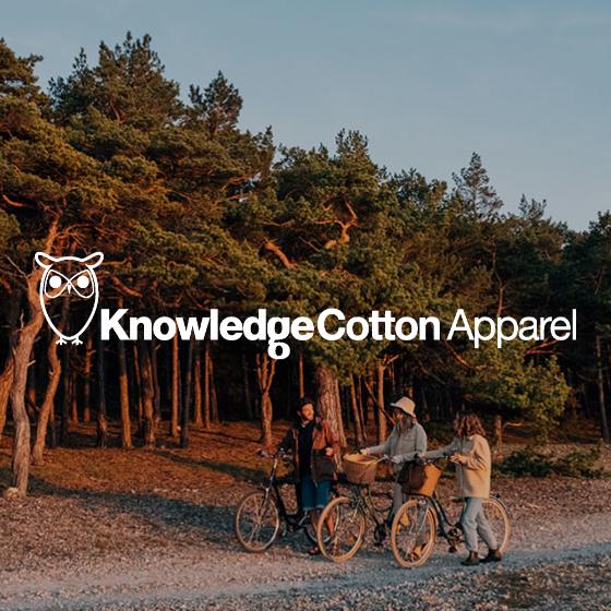 Drei Menschen mit Fahrrädern halten bei Sonnenuntergang auf einem Waldweg inne. Die Bäume sind üppig und der Himmel klar. Auf dem Bild ist das weiße Logo von „Knowledge Cotton Apparel“ mit einer Eule zu sehen.