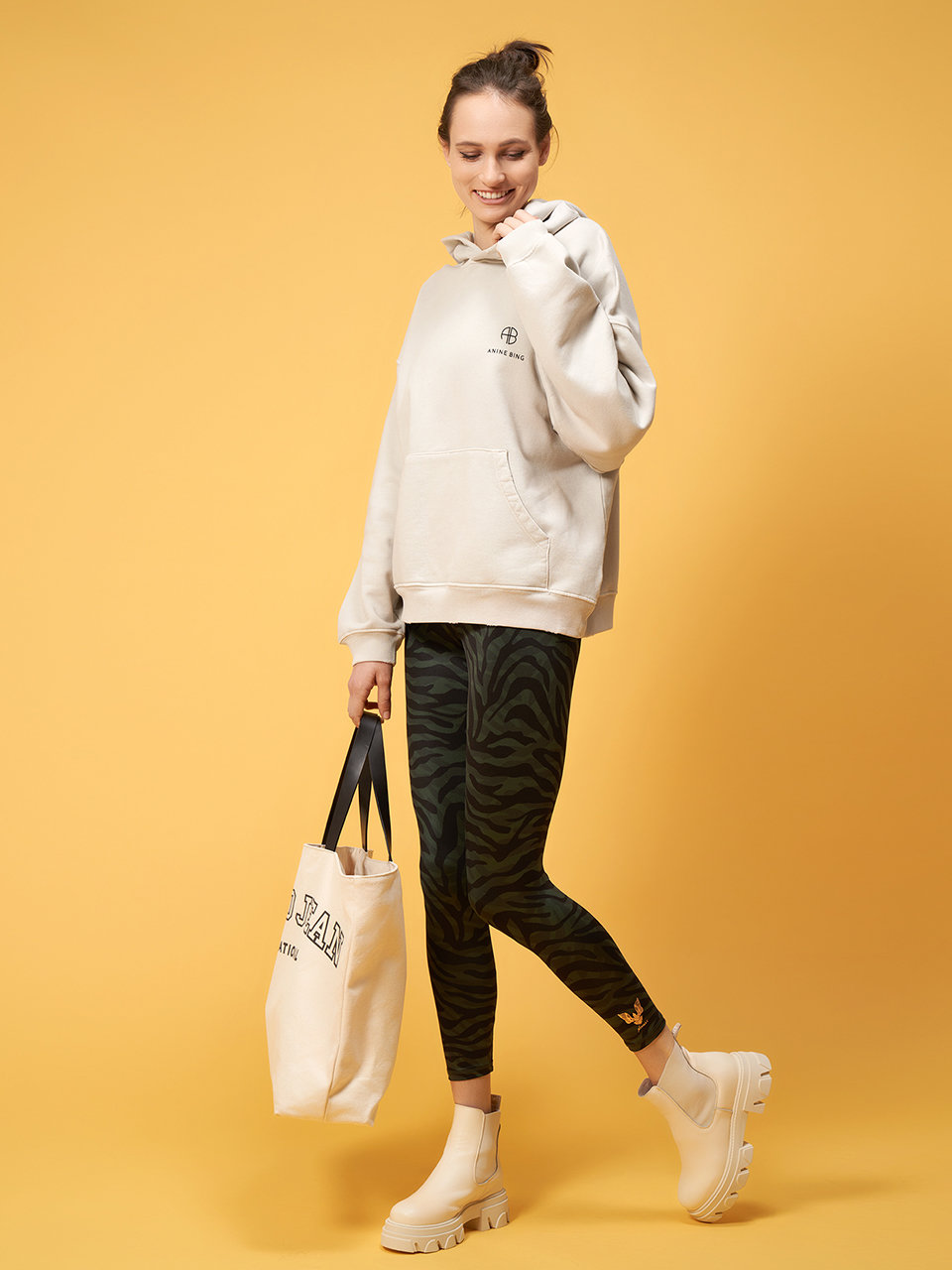 Eine Person in einem beigen Kapuzenpullover und Leggings mit Zebramuster posiert vor einem gelben Hintergrund. Sie hält eine Einkaufstasche und trägt cremefarbene Stiefeletten. Ihre Haare sind hochgebunden und sie lächelt mit einer Hand vor ihrem Gesicht.