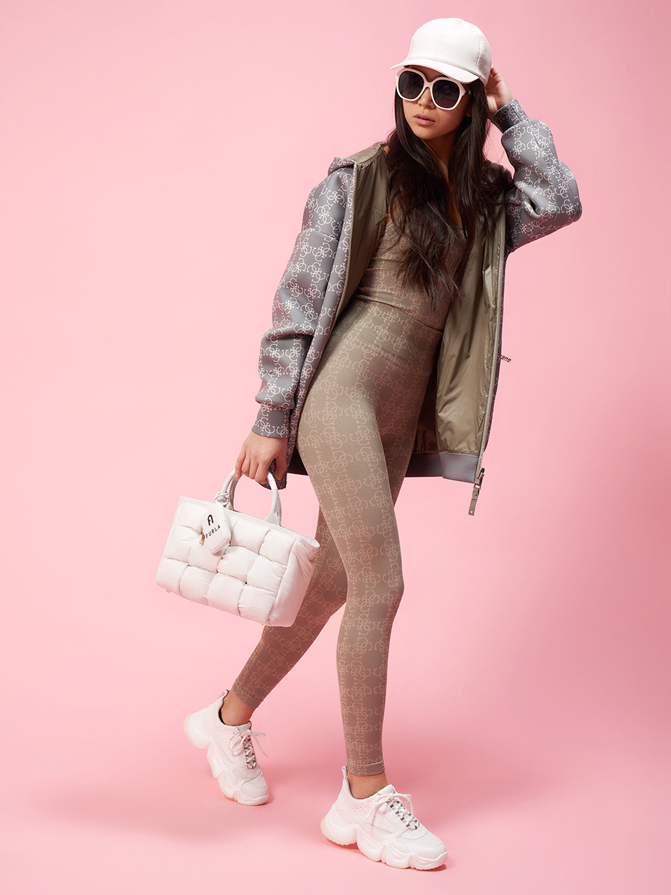 Eine Frau posiert vor einem rosa Hintergrund. Sie trägt eine grau gemusterte Jacke, passende Leggings, weiße Turnschuhe und eine weiße Mütze. Sie hält eine gesteppte weiße Handtasche und trägt eine Sonnenbrille. Sie strahlt einen sportlich-schicken Stil aus.