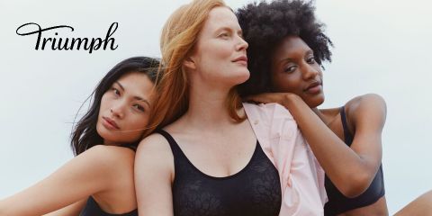 Drei Frauen mit unterschiedlichem Hautton und schwarzen und hautfarbenen BHs posieren eng beieinander und strahlen Selbstbewusstsein und Vielseitigkeit aus. Links ist der Text „Triumph“ in kursiver Schrift zu sehen. Der Himmel im Hintergrund ist hellblau.