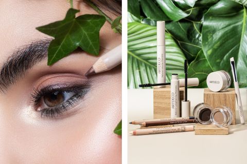 Nahaufnahme einer Person, die neben einer Auslage mit verschiedenen Make-up-Produkten von Artdeco, darunter Mascara, Augenbrauenstifte und Lidschatten, die auf Holzklötzen mit grünen Blättern im Hintergrund angeordnet sind, Eyeliner aufträgt.