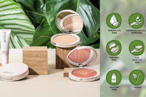Eine Präsentation von ARTDECO-Kosmetikprodukten mit einem natürlichen Concealer, zwei offenen Kompaktdosen mit Kompaktpuder und Rouge sowie Holzklötzen. Dahinter sind grüne Blätter und Symbole zu sehen, die Produkteigenschaften wie vegan und parabenfrei hervorheben.