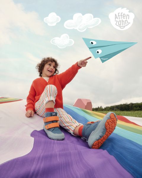 Ein Kind in einem leuchtend orangefarbenen Pullover und gestreiften Hosen sitzt auf einer bunten Fläche und zeigt auf ein Papierflugzeug im Comic-Stil am Himmel. Es lächelt und ist von flauschigen Wolken umgeben. In der Ecke steht „Affenzahn“.
