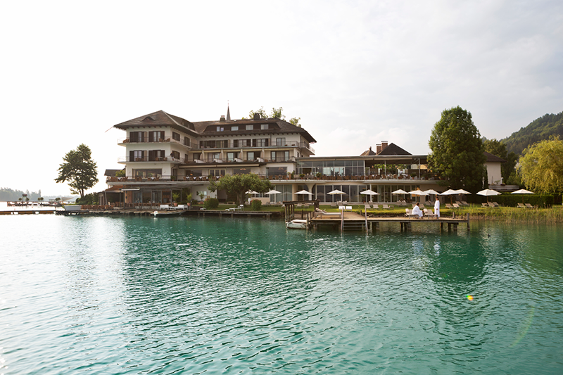 Ein großes Hotel am Seeufer mit mehreren Balkonen und Sitzgelegenheiten im Freien bietet Blick auf ein smaragdgrünes Gewässer. Sonnenschirme säumen ein Holzdeck und bieten schattige Sitzbereiche. Bäume und ein teilweise bewölkter Himmel rahmen die Szene ein.