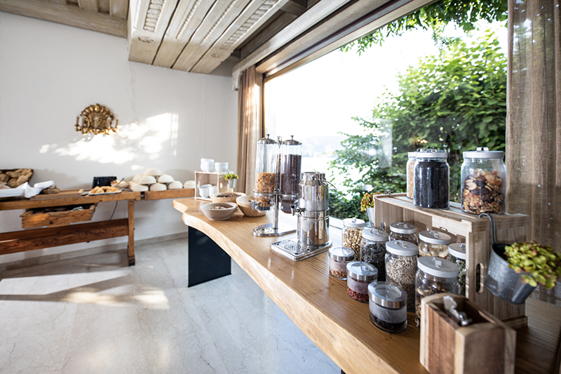 Ein heller Frühstücksbuffetraum mit einer Auswahl an Müsli, Marmelade und Brot auf Holztischen. Große Fenster lassen natürliches Licht herein und geben den Blick auf die grüne Umgebung frei. Das Ambiente ist elegant und einladend, mit ordentlich angeordneten transparenten Behältern.