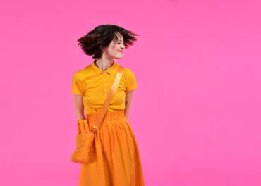 Eine Person mit kurzen Haaren und einem leuchtend gelben Kleid steht vor einem leuchtend rosa Hintergrund. Sie blickt mit einem leichten Lächeln zur Seite und ihre Haare sind mitten in der Bewegung eingefangen. Eine passende gelbe Tasche hängt über ihrer Schulter.