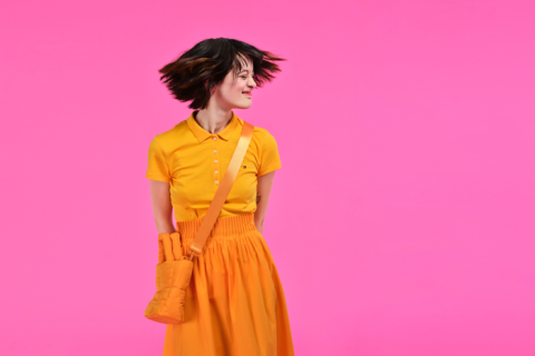 Eine Person mit kurzen Haaren und einem leuchtend gelben Kleid steht vor einem leuchtend rosa Hintergrund. Sie blickt mit einem leichten Lächeln zur Seite und ihre Haare sind mitten in der Bewegung eingefangen. Eine passende gelbe Tasche hängt über ihrer Schulter.