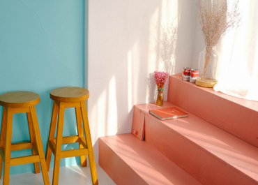 Ein minimalistisches Interieur besteht aus zwei Holzhockern vor einer türkisfarbenen Wand, rosa Stufen mit einem Buch, einer Vase mit rosa Blumen und Dosen auf den Stufen. Die Szene ist in warmes, natürliches Licht getaucht.