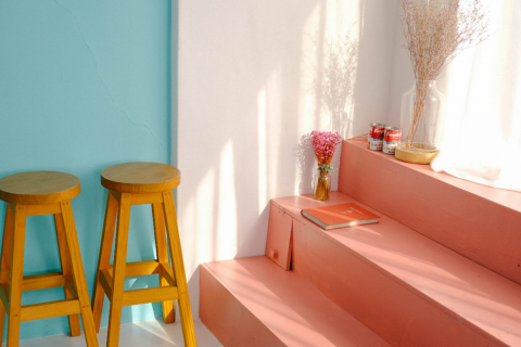 Ein minimalistisches Interieur besteht aus zwei Holzhockern vor einer türkisfarbenen Wand, rosa Stufen mit einem Buch, einer Vase mit rosa Blumen und Dosen auf den Stufen. Die Szene ist in warmes, natürliches Licht getaucht.