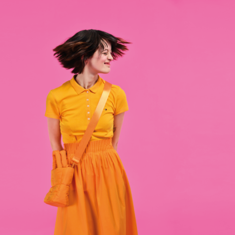 Eine Person in einem orangefarbenen Outfit mit passender Tasche steht vor einem leuchtend rosa Hintergrund, lächelt mit geschlossenen Augen und wehendem Haar.