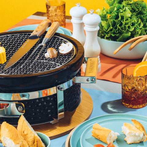 Ein Tisch mit einem kleinen Grill, Gemüse, Mais und Knoblauch darauf. Teller mit Brot und Salat stehen um den Grill herum. Zwei Gläser mit Orangensaft, Salz- und Pfefferstreuer und eine Schüssel mit Blattgemüse runden das Ambiente ab.