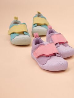 Zwei Paar Kleinkinderschuhe auf beigem Hintergrund: ein Paar ist helllila mit rosa Trägern, das andere hellblau mit gelben Trägern.