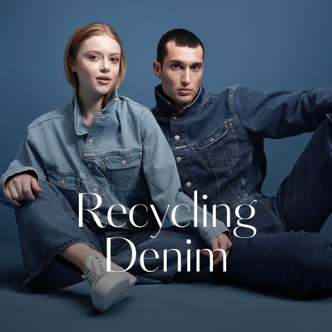 Zwei Personen in Jeans-Outfits posieren vor einem blauen Hintergrund mit dem Text "Recycling Denim" vor ihnen.