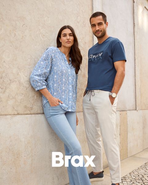 Eine Frau in blau gemusterter Bluse und heller Jeans steht neben einem Mann in blauem Brax-T-Shirt und beiger Hose. Sie lehnen an einer hellen Wand. Am unteren Bildrand ist das Wort „Brax“ zu sehen.