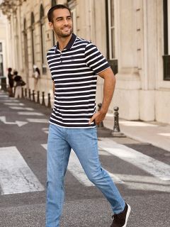 Ein Mann in einem schwarz-weiß gestreiften Poloshirt und hellblauen Jeans geht selbstbewusst über eine Straße. Im Hintergrund sind eine Reihe beigefarbener Gebäude und mehrere Fußgänger zu sehen.