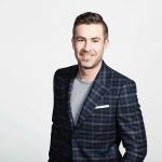 Ein Mann mit kurzen Haaren und Bart lächelt in die Kamera. Er trägt ein dunkel kariertes Jackett über einem grauen Hemd. Der Hintergrund ist schlicht weiß.
