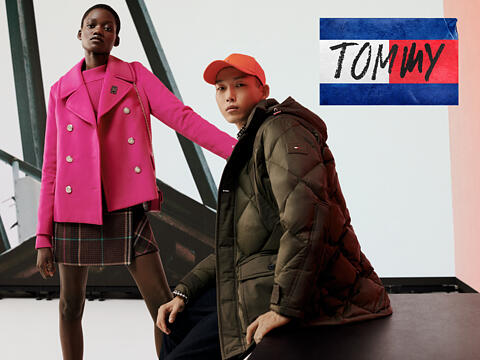 Ein Model in einer leuchtend rosa Jacke steht neben einem anderen Model in einer olivgrünen Pufferjacke und einer orangefarbenen Mütze. Im Hintergrund sind ein Bauwerk und das Tommy-Logo in Rot, Weiß und Blau zu sehen.