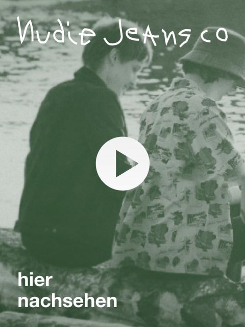 Zwei Personen sitzen im Profil am Wasser. Das Bild hat einen Grünstich und enthält oben den Text „Nudie Jeans Co“ und unten „hier nachsehen“, mit einem Play-Button-Symbol in der Mitte.