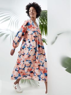 Eine Person lächelt, während sie ein langärmliges Kleid mit Blumenmuster trägt und neben großen grünen Blättern läuft. Das Kleid ist in den Farben Rosa, Blau und Weiß gehalten und sorgt so für einen lebendigen und sommerlichen Look. Sie trägt weiße Turnschuhe.